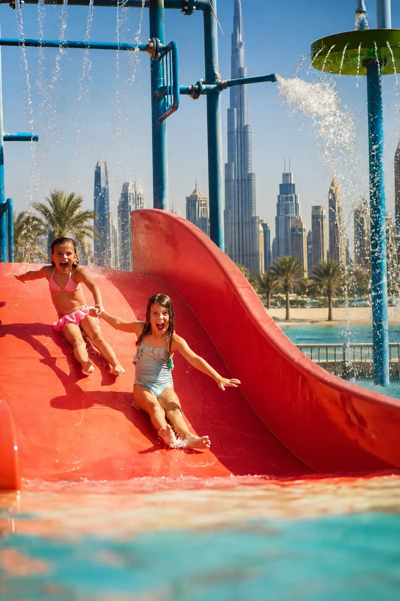 Two girls coming down a red water slide with the city skyline behind them